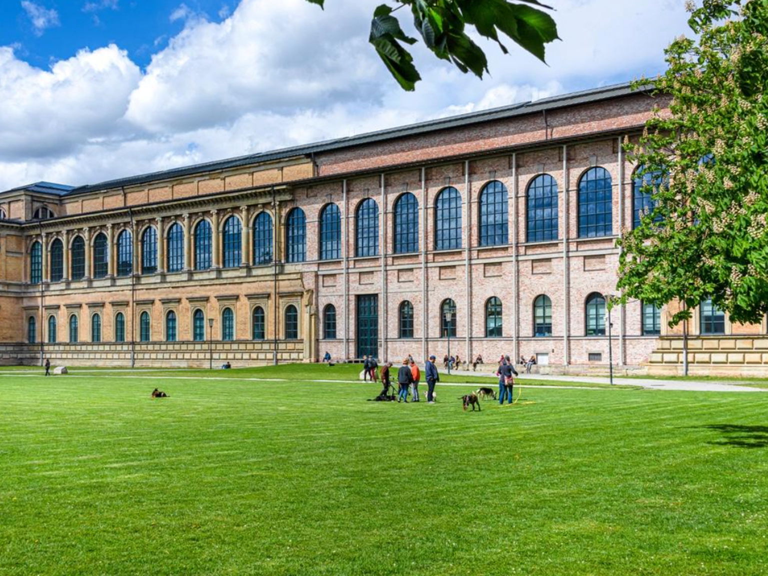 Brick university building with two stories of tall arched windows along a green lawn; people walking dogs in front.